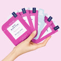 Woman’s hand holding Pedi in a Box Ultimate 6 Step Wild Black Plum packets on a light purple background.