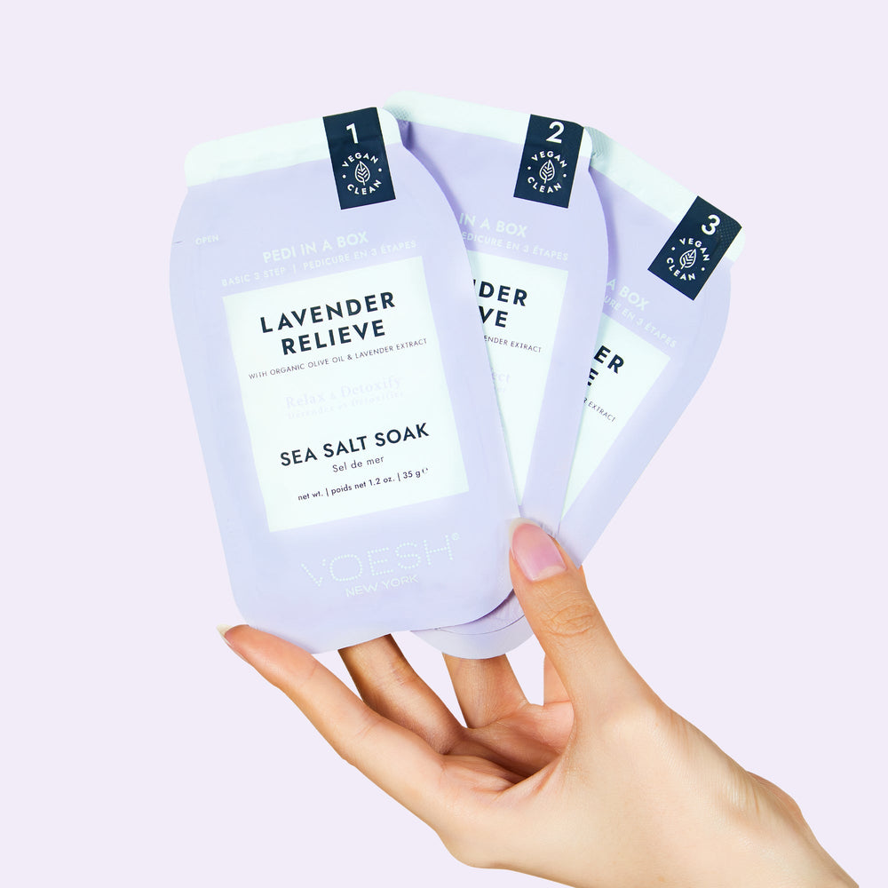 Woman's hand holding lavender relieve pedi in a box 3 step packets on light purple background