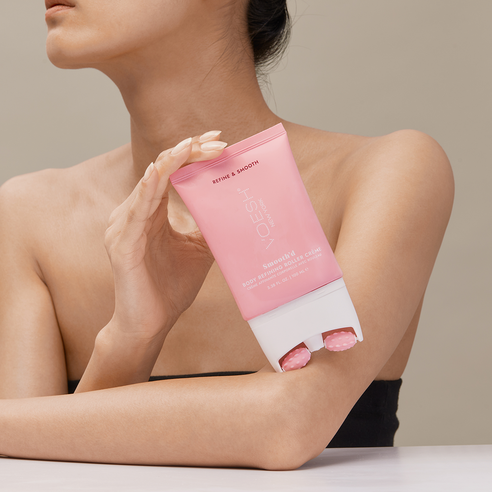 Woman holding Smooth’d Body Refining Roller Crème and leaning it on her forearm, pictured on a tan background.